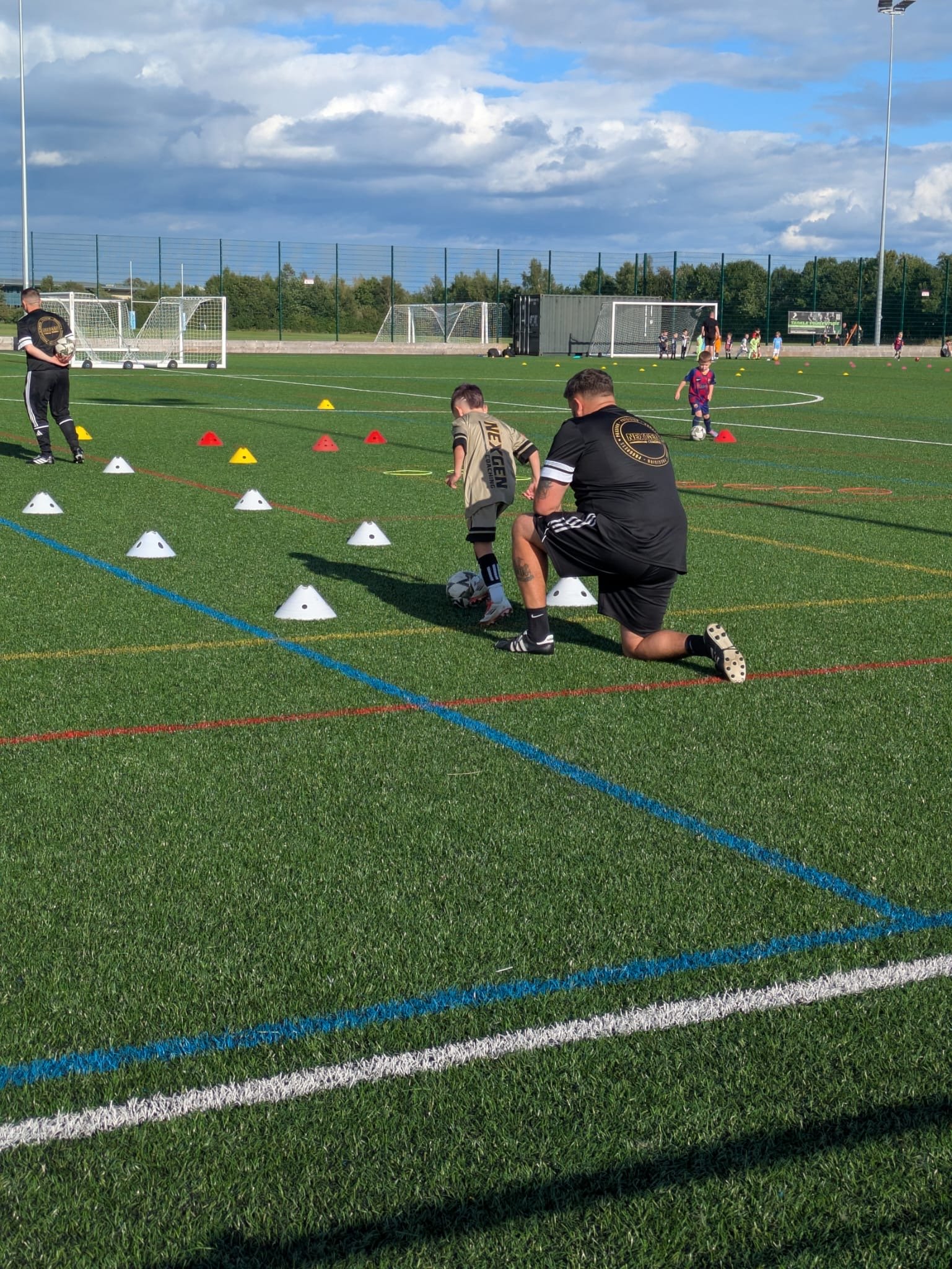 Nexgen coach kneeling beside a young player during a one-to-one session on an outdoor pitch