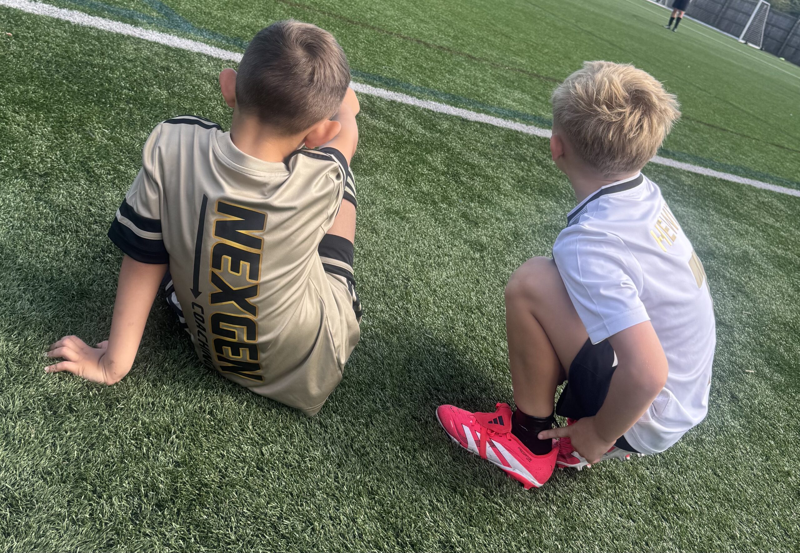 Two young players seen from behind, sitting on the pitch side by side, one in a Nexgen shirt