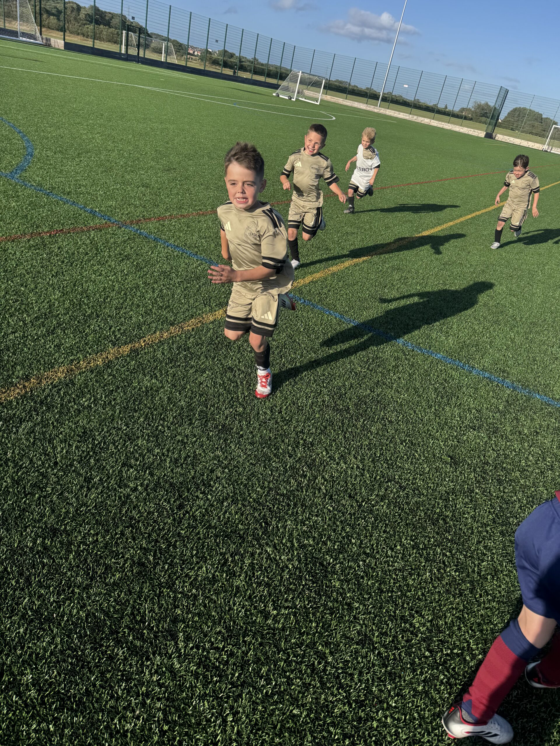 Young Nexgen players in gold training kit running with the ball on an outdoor 3G pitch