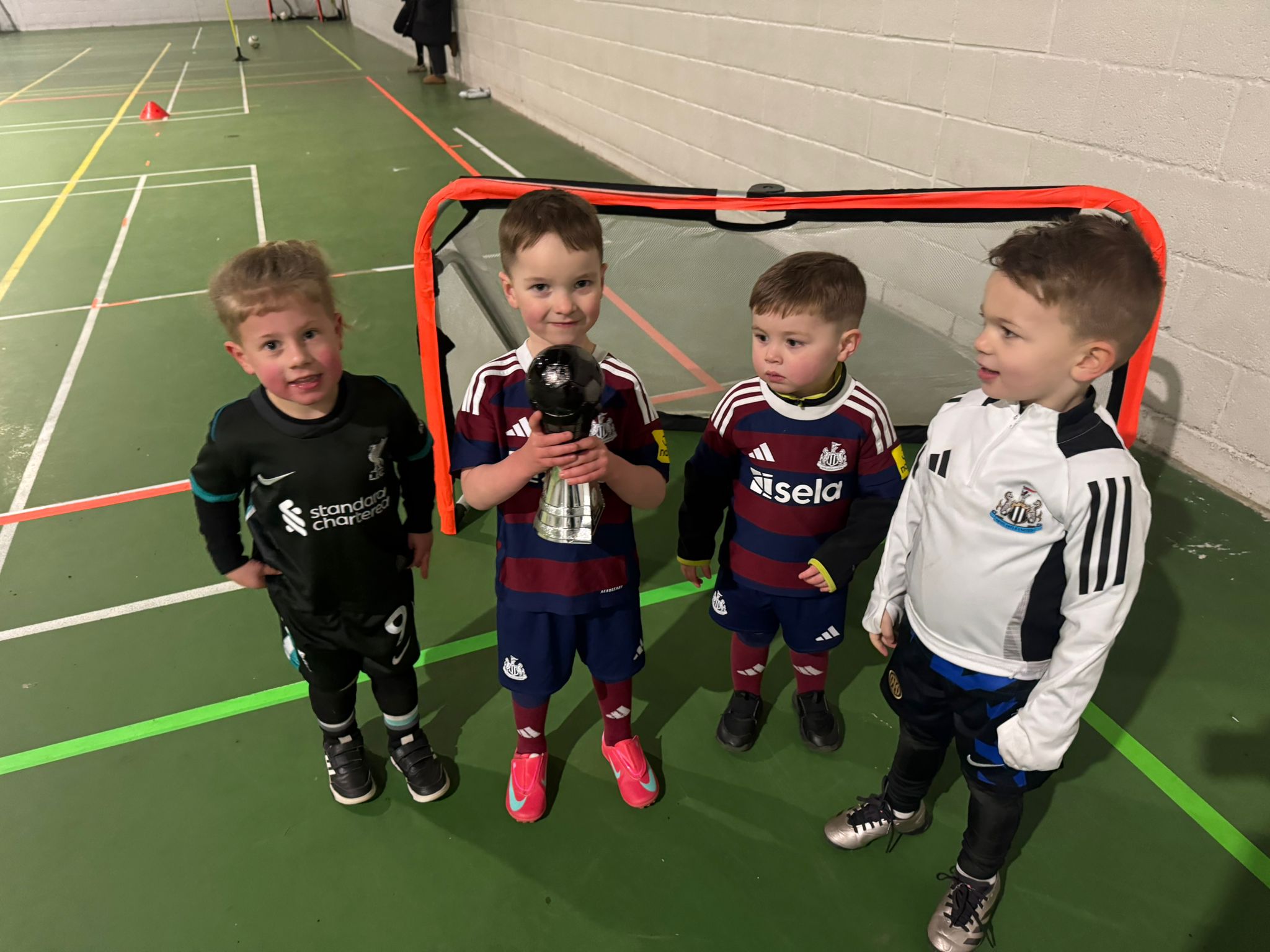 Four young players in football kit standing in front of a goal at an indoor small-group session